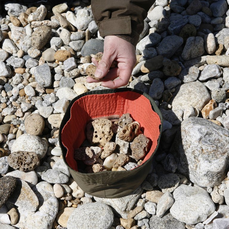 Eine Hand hebt Steine am Strand auf. Einer Touristin aus Belgien droht aktuell eine Haftstrafe, weil sie Steine aus dem Türkei-Urlaub mit nach Hause nehmen wollte. (Foto: IMAGO, robertharding)