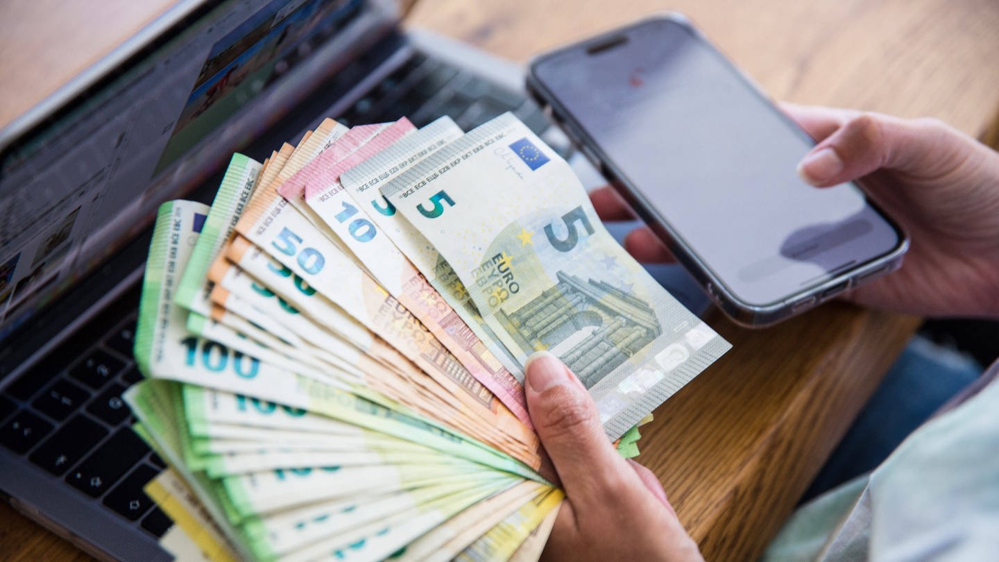 Eine Person hält mehrere tausend Euro in Bargeld in der Hand, dahinter steht ein Laptop und sie hält ihr Handy in der Hand. (Foto: IMAGO, IMAGO/Fotostand)