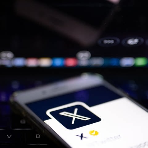 Auf dem offiziellen Profil der Plattform X auf dem Bildschirm eines Smartphones und auf dem Display eines Laptops ist der weiße Buchstabe X auf schwarzem Hintergrund zu sehen.