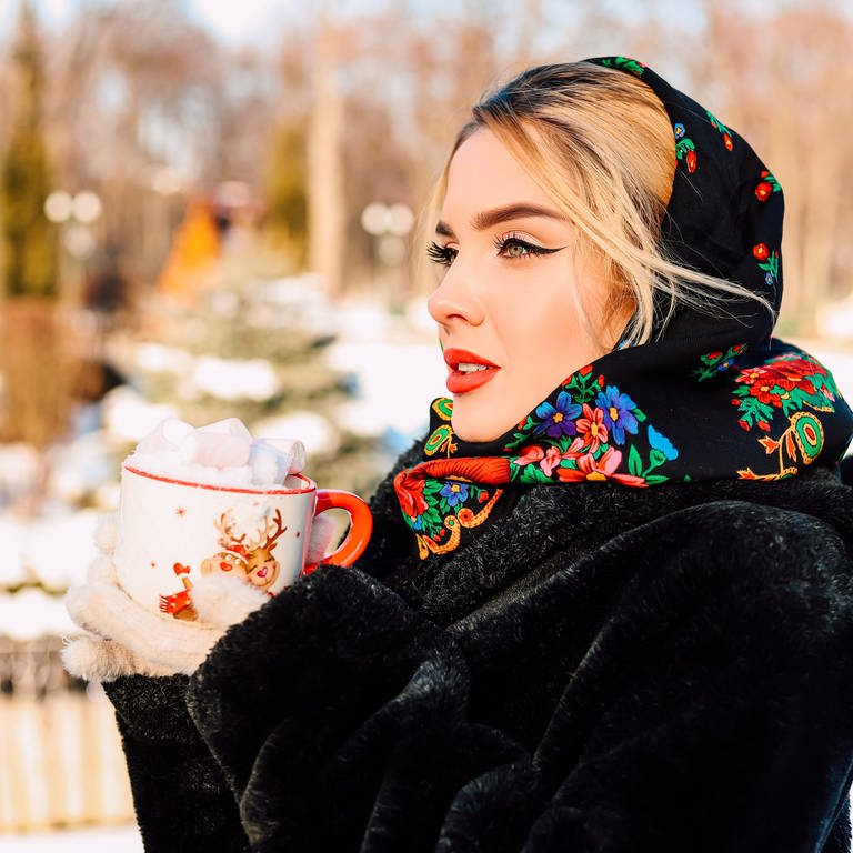SYMBOLBILD: Eine Frau steht draußen mit einer Tasse in der Hand im Schnee in einem Pelzmantel mit einem slavischen Tuch auf dem Kopf.