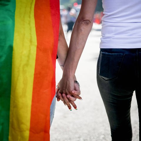 Zwei Personen halten Händchen und haben eine Regenbogen-Flagge umhängen.