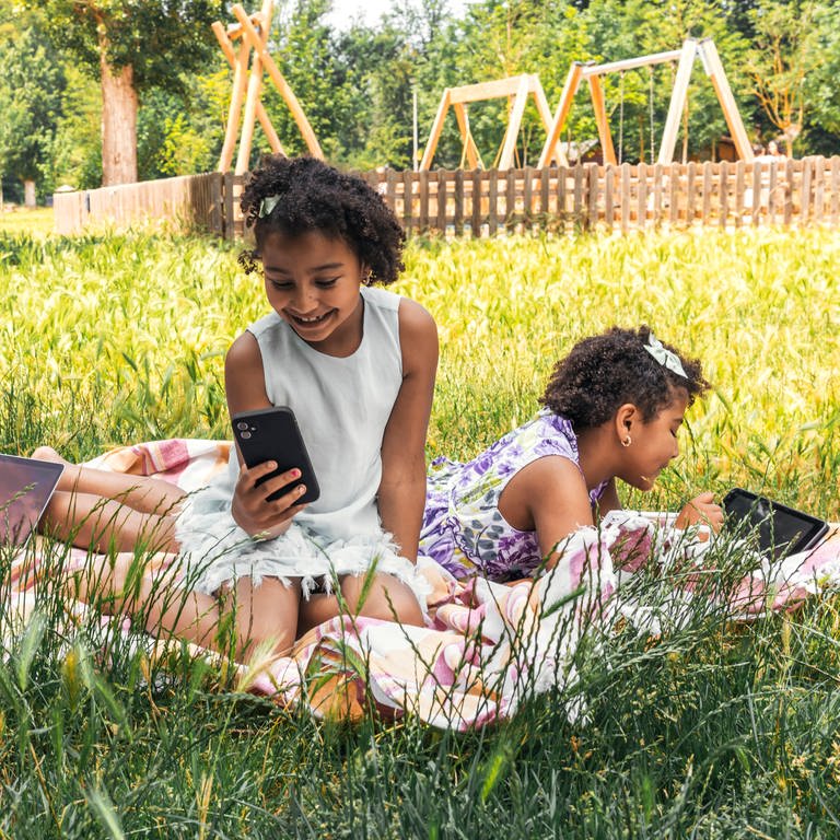 Glückliche Schwestern verbringen Zeit mit drahtloser Technologie im Gartenmodell (Symbolfoto). (Foto: IMAGO, Westend61)