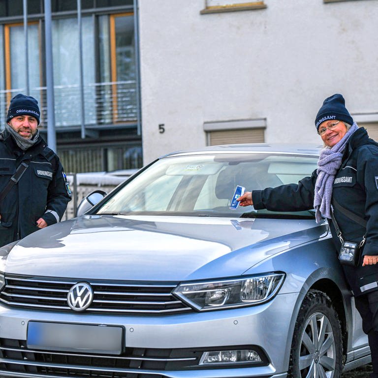 Zwei Mitarbeiter des Ordnungsamtes stehen auf einem Parkplatz in Giengen an der Brenz im Landkreis Heidenheim. Wer derzeit richtig parkt, bekommt Schokolade als Knöllchen.