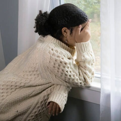 Depression: Eine junge Frau steht am Fenster und hält ihre Händen über das Gesicht (Symbolbild) (Foto: IMAGO, IMAGO / Cavan Images)