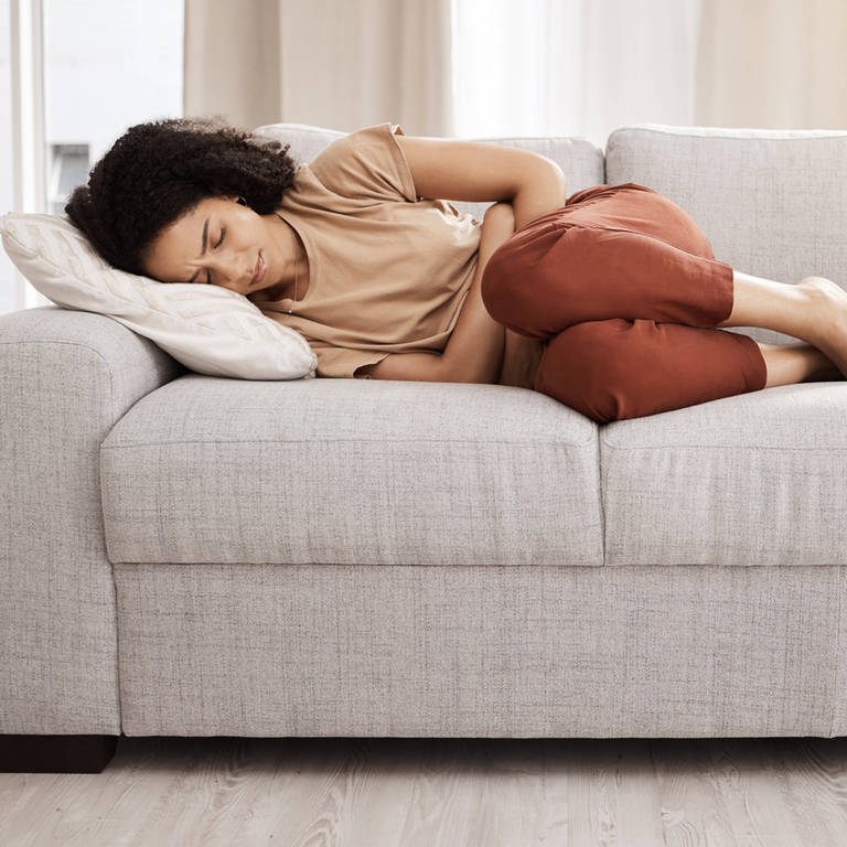Eine Frau liegt auf einer Couch und hält sich die Hände vor den Bauch wegen Unterleibsschmerzen.