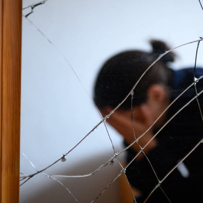 Eine Frau sitzt neben einem gesprungenen Spiegel auf einem Bett und hält ihren Kopf in den Händen (gestellte Szene)