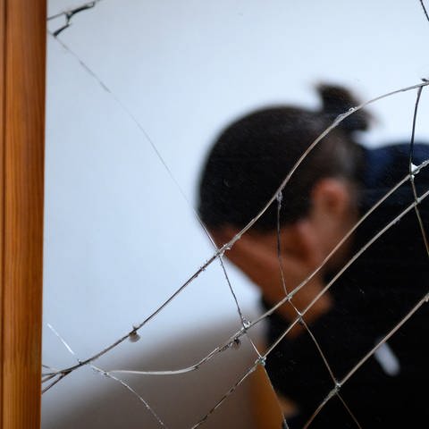 Eine Frau sitzt neben einem gesprungenen Spiegel auf einem Bett und hält ihren Kopf in den Händen (gestellte Szene) (Foto: dpa Bildfunk, picture alliance/dpa | Jonas Walzberg)