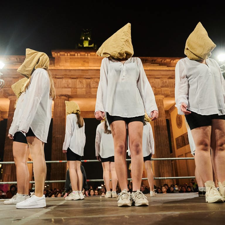 Tänzerinnen stehen mit Kopfbedeckungen auf einer Bühne vor dem Brandenburger Tor.