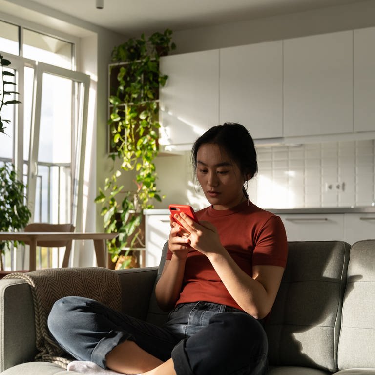 Eine junge Frau sitzt auf dem Sofa und guckt auf ihr Handy.