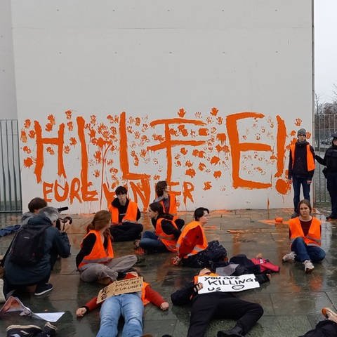 Aktivistinnen und Aktivisten der Letzten Generation sitzen vor einer Wand des Bundeskanzleramts auf der der Schriftzug «Hilfe! Eure Kinder» steht. Teilnehmer der Protestgruppe haben am Samstag die Fassade des Bundeskanzleramts mit orangener Farbe beschmiert.