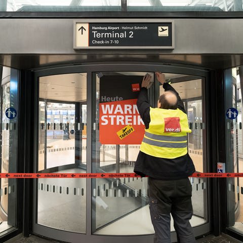 Ein Verdi-Vertreter hängt ein Plakat mit der Aufschrift «Warnstreik» an einer geschlossenen Terminaldrehtür am Hamburger Flughafen auf. (Foto: dpa Bildfunk, picture alliance/dpa | Bodo Marks)