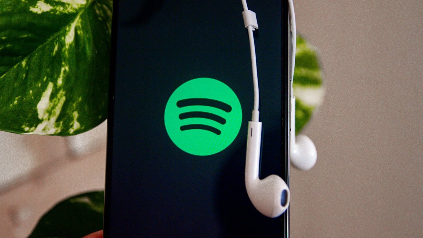 Ein Handy zeigt das Spotify-Logo. Über dem Gerät hängen Kopfhörer. (Foto: IMAGO, IMAGO / Ato Press)