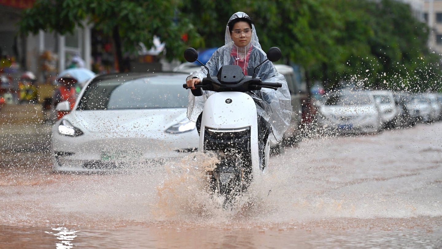 Eine Frau fährt mit ihrem Roller über eine überflutete Straße. (Foto: IMAGO, IMAGO / Xinhua)