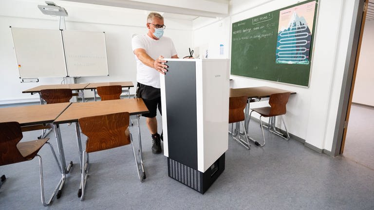 Ein Hausmeister schiebt einen recht großen Luftfilter durch ein Klassenzimmer. (Foto: dpa Bildfunk, picture alliance/dpa | Julian Stratenschulte)